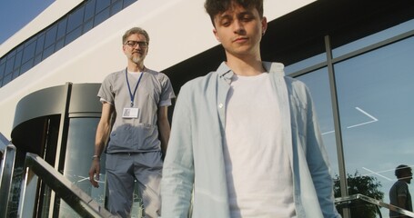 Mature doctor with tablet in hands stands with patient at clinic entrance, discuss treatment. Healthcare specialist says goodbye to client after appointment, then looks at camera. Medic works outside.