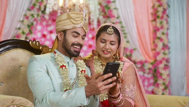 Happy Indian newlywed couples using or checking mes messages or wishes on mobile phone on wedding stage - concept of social media sharing, Marriage ceremony and affection