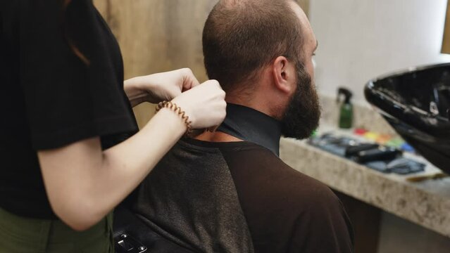 The photo shows a young stylish female hairdresser putting a cape on a client. Fashionable hairdresser at work in a retro barbershop. Preparing for a haircut and grooming for a stylish man.