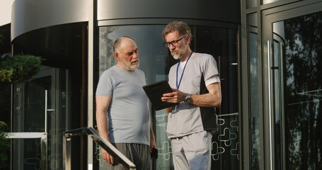 Mature physician with digital tablet in hands stands with aged patient at clinic entrance and discuss his treatment. Health care specialist consults and supports elderly client. Medic works outside.