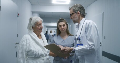 Mature doctor walks the clinic corridor, comes to colleague and elderly patient. Physician shows test results on papers to nurse and woman. Medical staff and patients in hospital hallway.