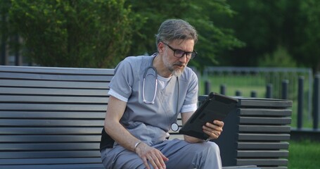 Obraz premium Doctor in uniform sits on bench outside modern hospital, talks to patient about treatment during online appointment using digital tablet. Medical staff of clinic or medical center. Healthcare system.