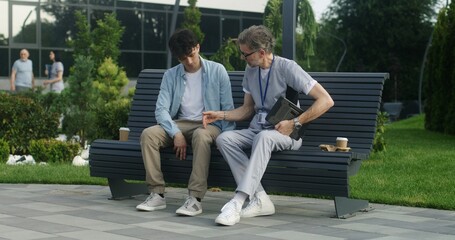 Mature doctor with tablet in hands sits with middle aged patient on bench and consults him. Man...