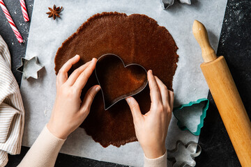 baking, cooking and christmas concept - close up of hands with mold cutting gingerbread dough on black kitchen table top