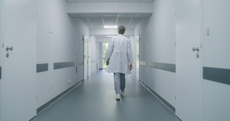 Mature doctor walks down hospital corridor with digital tablet computer to medical room. Professional medic stops to talk to young colleague and old patient. Medical staff at work in medical center.