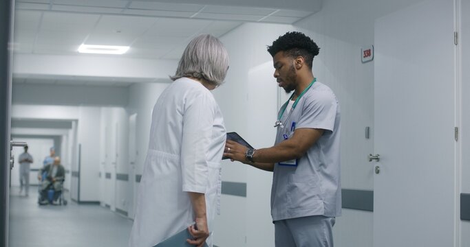 African American doctor uses digital tablet computer, shows MRI or CT scanning results to female colleague. Multicultural medics discuss medical test results of patient in hospital or clinic corridor.
