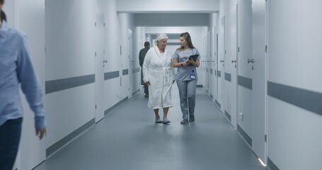 Female medic, nurse with digital tablet computer and elderly woman talk, walk along the clinic corridor to hospital room after procedures. Medical staff, doctors, patients in medical center hallway.