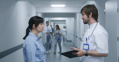 Male doctor stands in clinic corridor with female patient, uses digital tablet. Professional medic talks to adult woman, discusses medical tests results. Medical staff and patients in hospital hallway
