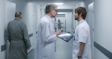 Obraz premium Mature doctor shows MRI, CT brain scan image to colleague using digital tablet. Professional medics stand and walk along the clinic corridor, discuss work. Medical staff, patients in hospital hallway.