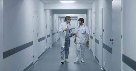 Medics walk along the clinic corridor, discuss work. Mature doctor shows MRI, CT brain scan image to colleague using digital tablet. Medical staff and patients in hospital or medical center hallway.