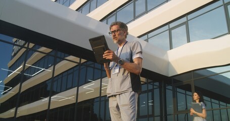 Doctor consults patient during online appointment using digital tablet. Medic in uniform stands outdoors near hospital. Medical staff walk in background. Clinic or medical center. Healthcare system.