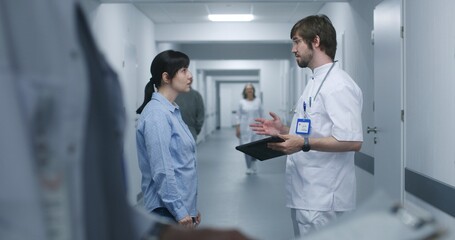 Male doctor stands in clinic corridor with female patient, uses digital tablet. Professional medic talks to adult woman, discusses medical tests results. Medical staff and patients in hospital hallway