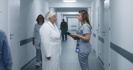 Female doctor, nurse with digital tablet and patient talk. Medic stands in modern clinic corridor with elderly woman after procedures. Medical staff and patients in hospital or medical center hallway.