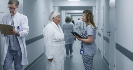 Female doctor, nurse with digital tablet and patient talk. Medic stands in modern clinic corridor with elderly woman after procedures. Medical staff and patients in hospital or medical center hallway.