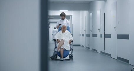 Male doctor stands with elderly woman in wheelchair in clinic corridor, uses mobile phone. Medic with female patient near operating or procedures room. Medical staff and patients in hospital hallway.