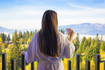 Brunette girl long hair from back in bathrobe standing on balcony. Elegant woman is drinking tea or coffee from a white cup enjoying morning. - obrazy, fototapety, plakaty