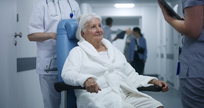 Elderly woman in wheelchair talks to doctor in clinic corridor. Medic discusses tests results with patient, uses digital tablet. Medical staff and patients walk in background in hospital hallway.