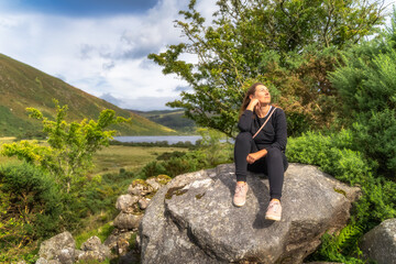 Naklejka na ściany i meble Young woman sitting on a large rock and smiling. Lake Lough Dan and mountain valley in background, Wicklow Mountains, Ireland