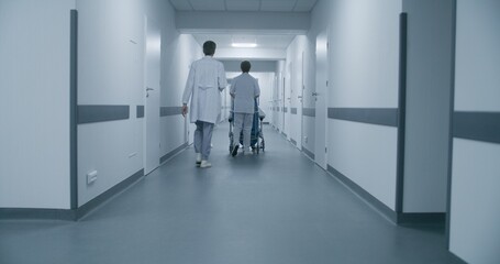 Back view of doctors pushing transfer wheelchair with patient walking along corridor. Medics talk, take person on procedures, therapy or to hospital room. Medical staff and patients in clinic hallway.