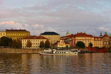 Cityscape with historic old buildings with spires and red roofs, and trees on the riverbank with boats. Autumn, late evening, sunset. Prague, Czech Republic, Vltava River, October 2022