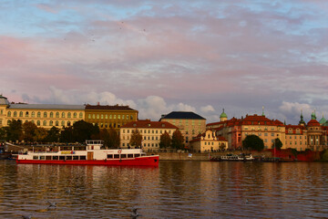 Cityscape with historic old buildings with spires and red roofs, and trees on the riverbank with red boat. Autumn, late evening, sunset. Prague, Czech Republic, Vltava River, October 2022