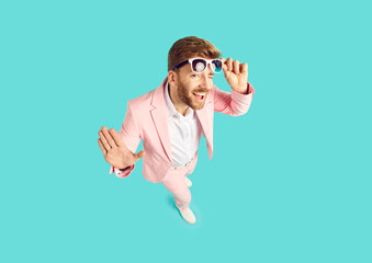 High angle shot from above of happy cheeky handsome young man wearing pink suit standing on blue color background looking at camera, smiling, taking off sunglasses and winking his eye. Fashion concept