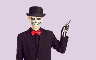 Portrait of spooky man with skull makeup pointing his finger to side. Handsome man wearing bowler hat, black suit and bow tie standing on isolated studio background. Day of Dead, Halloween concept