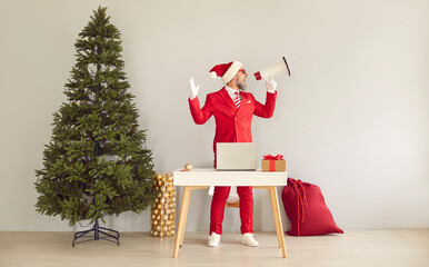 Obraz premium Funny excited man in red suit and Santa Claus cap standing by table near wall in office room with big green Christmas tree and red gift bag, advertising online holiday sale, yelling through megaphone
