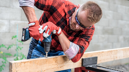 Cordless drill in worker hand while drilling a hole in the wooden batten. - obrazy, fototapety, plakaty