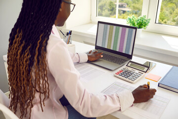 Young African American woman accountant sitting at office desk, using calculator, typing on notebook laptop PC computer, working with digital payroll document spreadsheets and taking notes on paper - obrazy, fototapety, plakaty