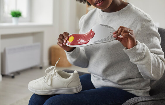 Cropped shot of young African American woman with shoe on her lap sitting on chair at home and looking at orthotic arch support insole that she is holding in her hands. Footwear, feet health concept