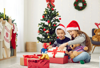Happy little children opening presents on Christmas morning. Two excited kids in red hats sitting on floor in beautiful, decorated living room and together looking inside box with wonderful Xmas gift