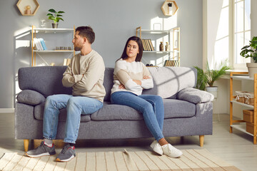 Family couple having conflict. Young man and woman quarrel and don't talk to each other. Two angry people sit down on sofa, turn away from each other, and keep sitting back to back, ignoring one other - obrazy, fototapety, plakaty