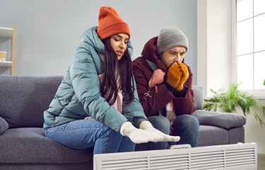 Young couple is sitting on the sofa at home in warm jacket and hat, warming their hands with heater...