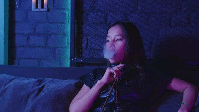 Asian young woman takes of mouthpiece to her lips, inhales and blows out puffs of white smoke at shisha cafe. Pretty adult girl smoking hookah sitting at couch at bar. Female smoker, bad habit