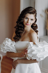Fototapeta premium Beautiful model-looking girl with long curly hair in a fashionable off-the-shoulder wedding dress. Vertical close-up portrait