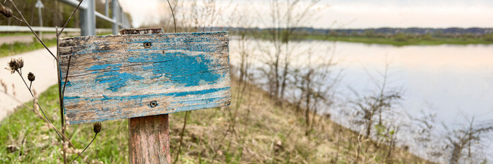 Wooden sign with peeling paint. A sign with old blue paint is embedded in the ground near the river and garden.