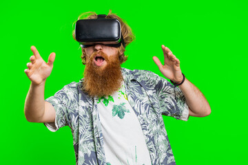 Excited happy bearded man using headset helmet app to play simulation game. Watching virtual reality 3D 360 video. Redhead guy in VR goggles isolated on green chroma key background. Future technology