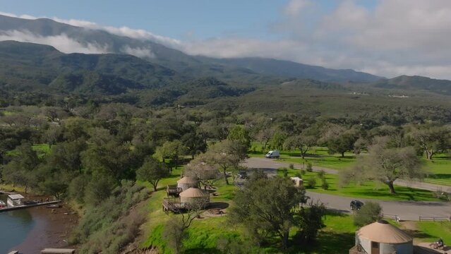 Forwards fly above campground at lake. Yurts, trees and green lawn. Cars passing on roads. High mountains in background. Cachuma Lake, California, USA