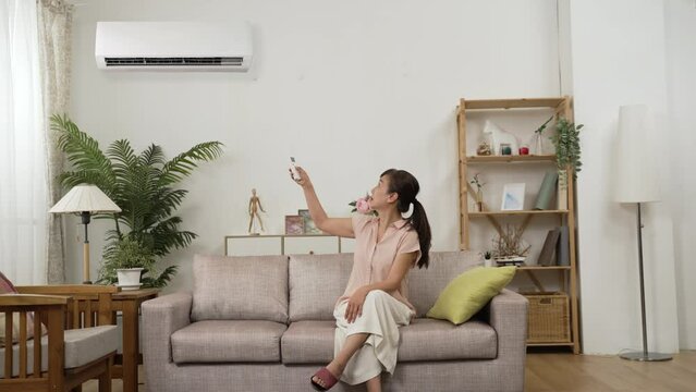 sweating asian mature woman feeling dehydration during menopause and fanning with hands. she switches on air conditioner and enjoys cool air with spreading arms on sofa at home