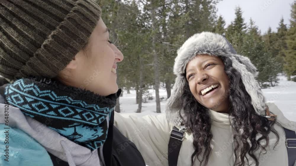 Wall mural Two beautiful smiling girlfriends enjoy winter hiking day in snow mountain. Multiracial cheerful female couple laughing together in nature. Young women look at each other cheerful on forest vacation.