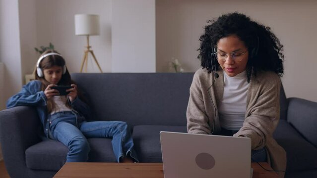Young adult woman with headset working on laptop computer sitting on sofa with her child using mobile phone at home. Family conciliation, working at home and technology lifestyle concept.