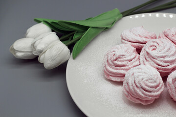 Delicate handmade pink marshmallows on a white plate with white tulips on a gray background.
