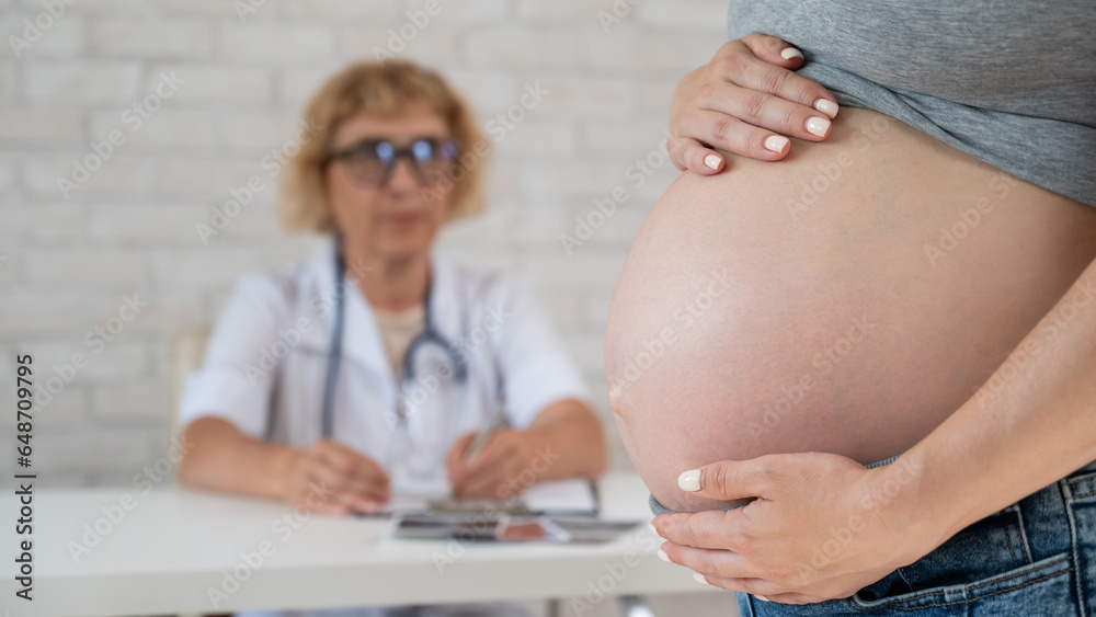 Wall mural Doctor obstetrician gynecologist at his desk in the background. Close-up of a pregnant woman's belly.