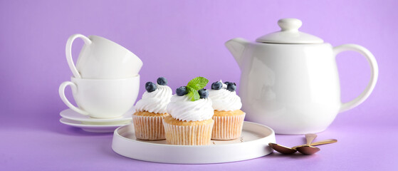 Tasty blueberry cupcakes, teapot and cups on lilac background