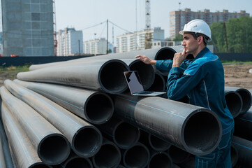 Caucasian male builder in a hard hat stands near the pipes and uses a laptop at a construction...
