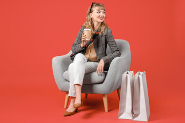 Stylish young woman with coffee cup and shopping bags in armchair on red background