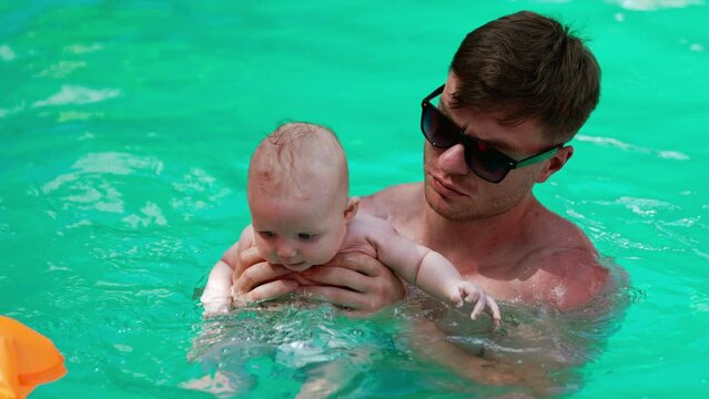 Tiny infant in a diaper is taught to swim in the pool by his dad. Family with a baby on vacation in summer.