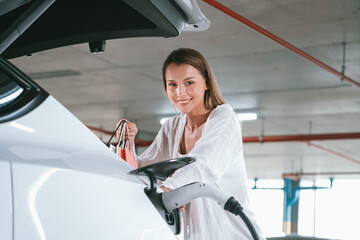 Young woman travel with EV electric car to shopping center parking lot charging in downtown city...