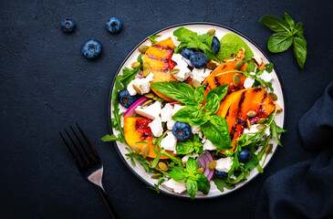 Gourmet salad with grilled peaches, blueberries, red onion, feta cheese, basil and mixed herbs, black table background, top view